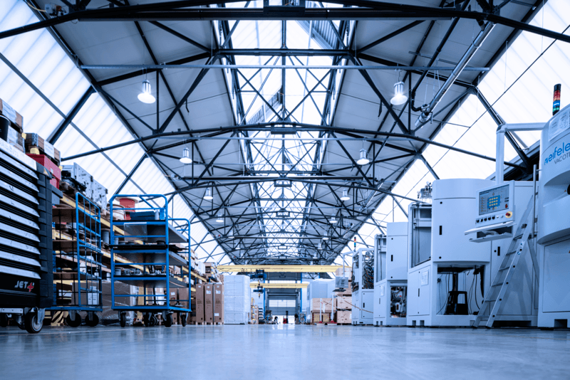 A spacious, modern factory interior of Eifeler with high ceilings, industrial lighting, shelves of supplies, carts, and large machinery, all organized in a clean, well-lit environment.