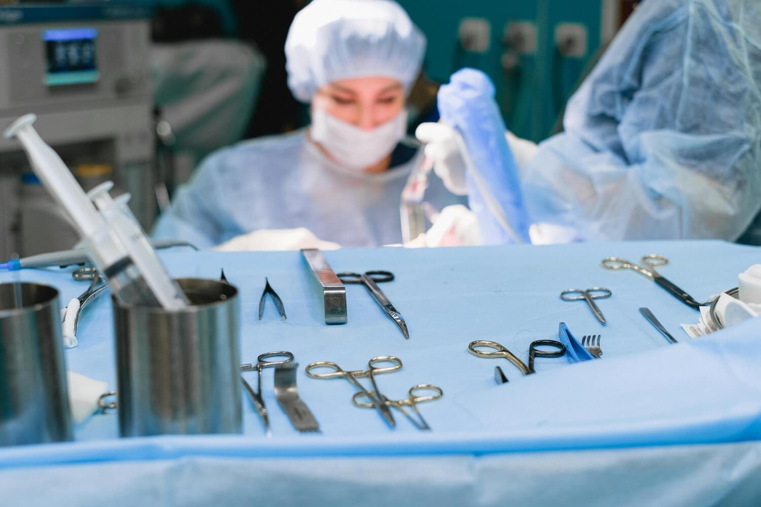 A tray with various surgical instruments is set up in the foreground, while blurred medical professionals in scrubs and masks work in the background.