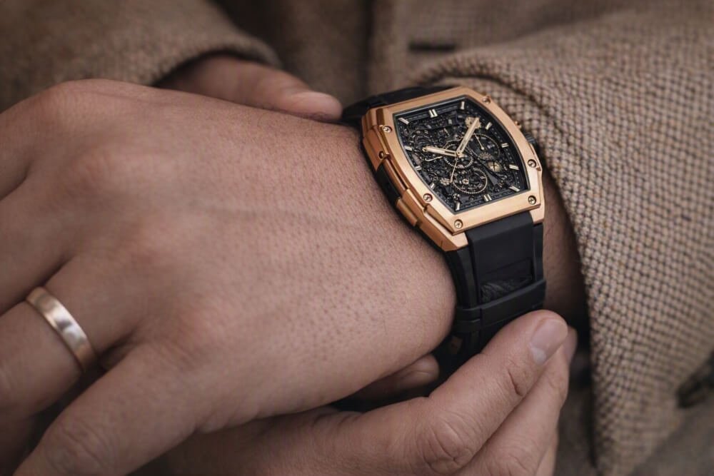 A person wearing a gold, square-faced wristwatch with a black strap and intricate dial, dressed in a brown textured coat, and a simple gold ring on one finger.