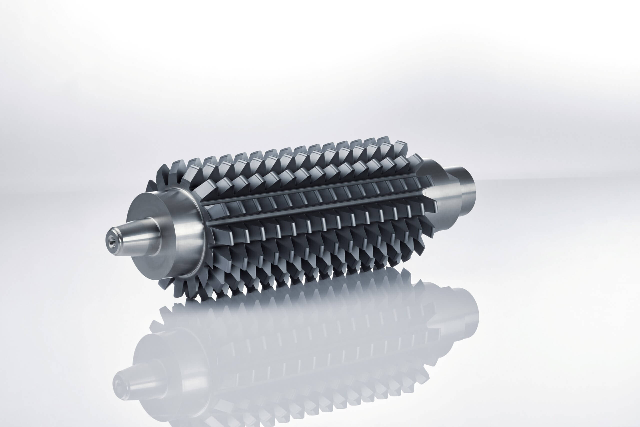 A large cylindrical metal gear with multiple angled teeth and smooth end shafts, placed on a reflective surface against a light gray gradient background.